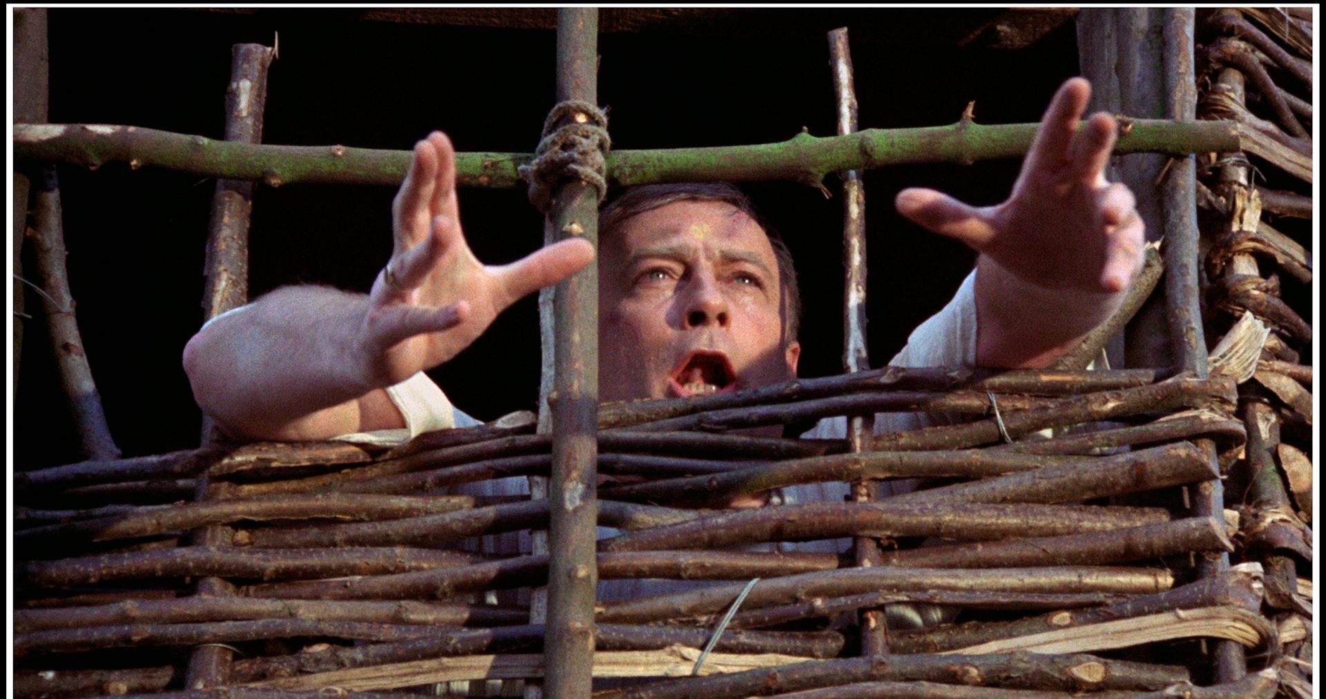 A man imprisoned in a makeshift prison made of wood, his hands outstretched in plea.